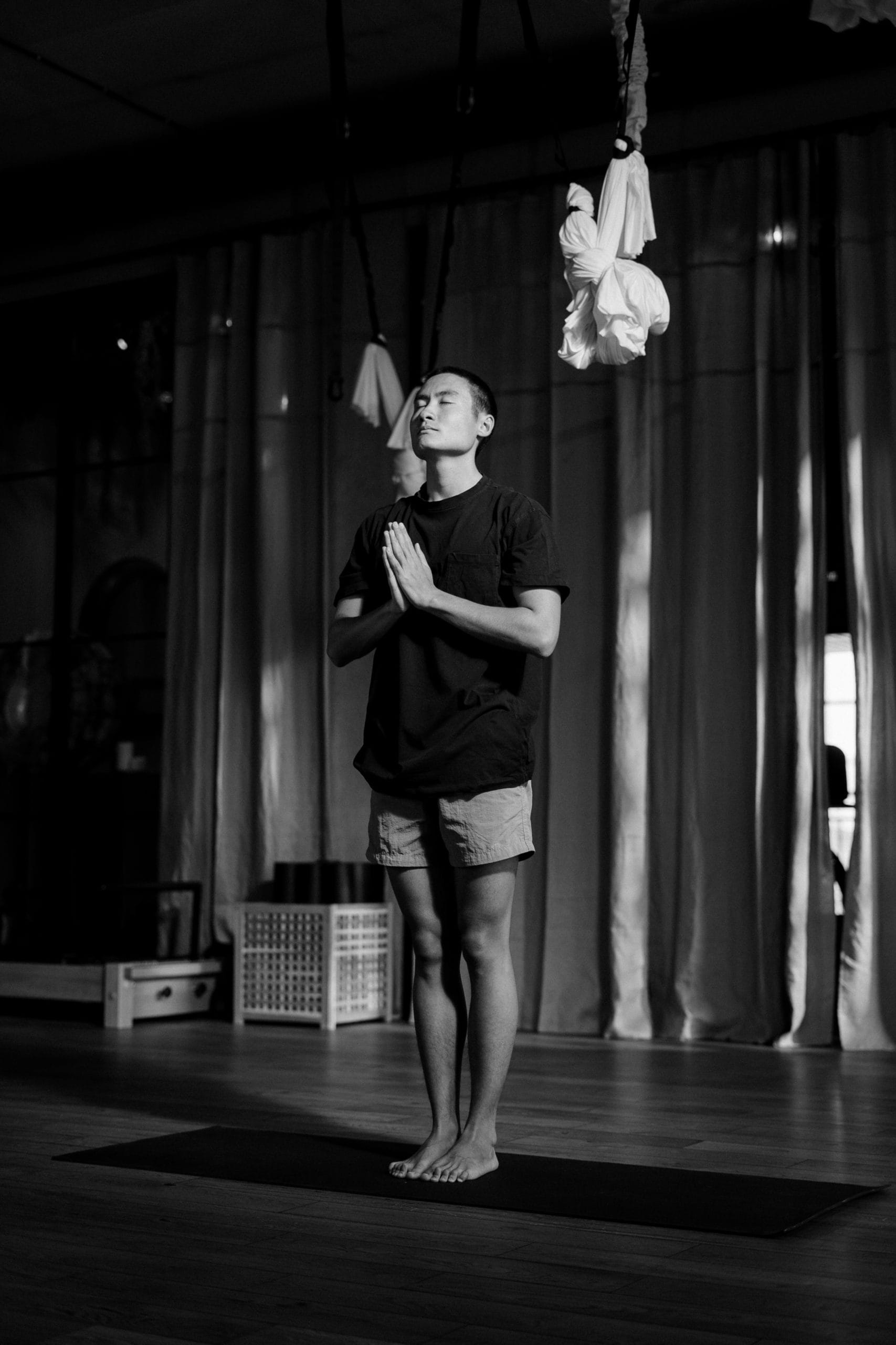 man with short hair wearing t shirt and shorts meditating to keep a positive mindset standing on mat in gym with hands clasped in front of him